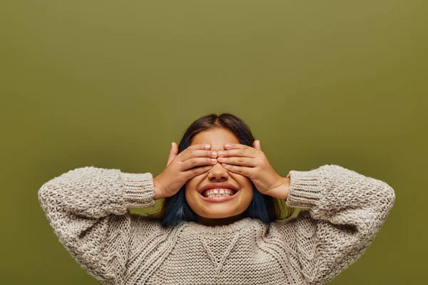 Joyeux preteen et fille élégante avec les cheveux teints portant un pull tricoté et couvrant les yeux avec les mains tout en se tenant isolé sur vert, fashion-forward preteen avec le sens du style — Photo de stock