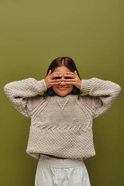 Smiling preteen child with colored hair wearing stylish knitted sweater while covering face with hands and standing isolated on green, fashion-forward preteen with sense of style — Stock Photo