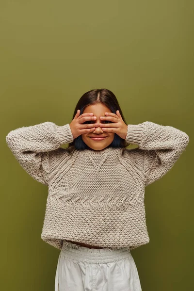 Enfant préadolescent souriant avec des cheveux colorés portant un pull tricoté élégant tout en couvrant le visage avec les mains et debout isolé sur vert, préadolescent avant de la mode avec un sens du style — Photo de stock