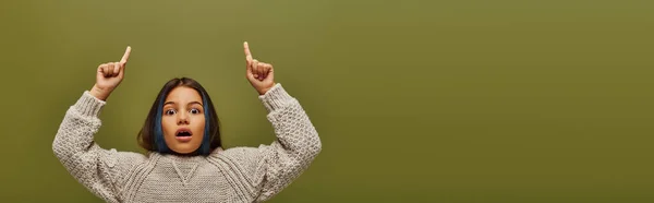 Impresionado niño preadolescente con el pelo de color que lleva suéter de punto moderno mientras mira a la cámara y señala con los dedos aislados en verde, pancarta, preadolescente de moda con sentido del estilo - foto de stock