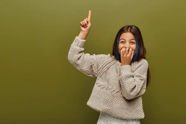 Laughing and stylish preteen girl with dyed hair wearing knitted sweater and pointing with finger while standing and posing isolated on green, fashion-forward preteen with sense of style — Stock Photo