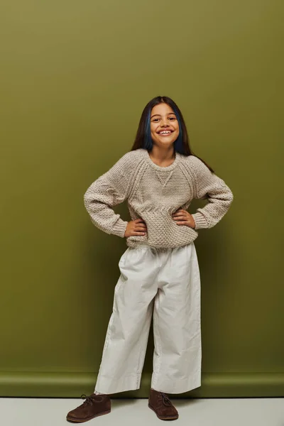 Full length of cheerful and stylish preteen girl with colored hair wearing knitted sweater and looking at camera while posing on green background, fashion-forward preteen with sense of style — Stock Photo