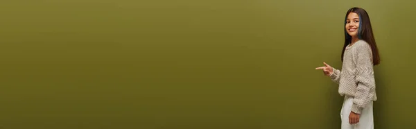 Chica preadolescente alegre en suéter de punto acogedor moderno apuntando con el dedo y mirando a la cámara mientras está de pie sobre fondo verde, pancarta con espacio de copia, moda preadolescente moderna y moderna - foto de stock