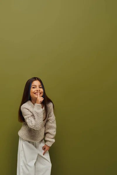 Chica preadolescente alegre y elegante con el pelo teñido en suéter de punto mostrando gesto secreto y mirando a la cámara mientras está de pie sobre fondo verde, moda preadolescente moderna y moderna - foto de stock
