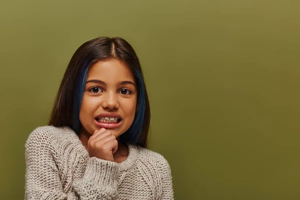 Retrato de niña preadolescente disgustada con el pelo de color usando suéter de punto acogedor y mirando a la cámara mientras está de pie y posando aislado en verde, moda preadolescente moderna - foto de stock