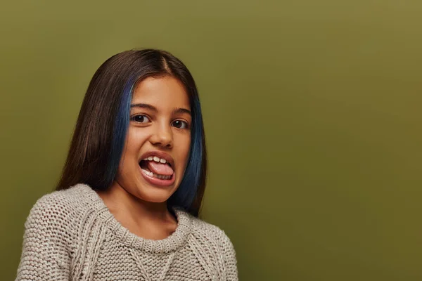 Portrait of stylish preteen girl with dyed hair wearing knitted sweater and sticking out tongue while looking at camera isolated on green, modern and hip preteen fashion concept — Stock Photo