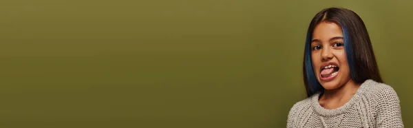 Retrato de la chica preadolescente de moda con el pelo teñido usando suéter de punto mientras que sobresale la lengua y mirando a la cámara aislada en verde, banner, moderno concepto de moda preadolescente - foto de stock