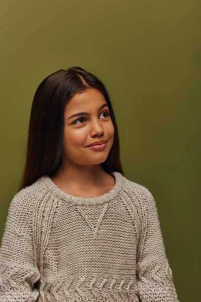 Portrait of pensive and stylish brunette preteen girl in knitted sweater looking away while standing and posing isolated on green, stylish girl in cozy fall attire concept — Stock Photo