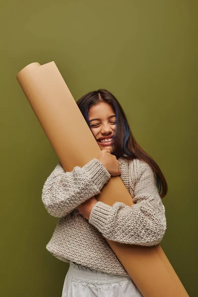 Pleased and stylish preadolescent girl with dyed hair wearing knitted sweater while hugging rolled paper and standing isolated on green, girl radiating autumn vibes concept — Stock Photo