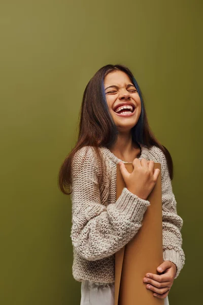 Веселая и стильная девочка-подросток в современном трикотажном свитере и осеннем наряде, держа в руках бумагу и смеясь стоя изолированно на зеленом, девушка излучает осенние вибрации концепции — стоковое фото
