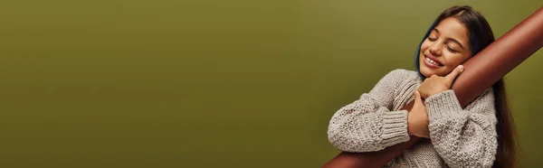 Menina pré-adolescente alegre e elegante com cabelo tingido vestindo camisola de malha enquanto abraçando papel laminado e de pé isolado em verde, menina irradiando vibrações de outono, banner com espaço de cópia — Fotografia de Stock