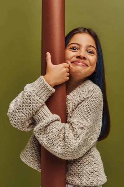 Portrait of excited and smiling trendy preteen girl in knitted sweater hugging rolled paper and looking at camera while standing isolated on green, girl radiating autumn vibes concept — Stock Photo