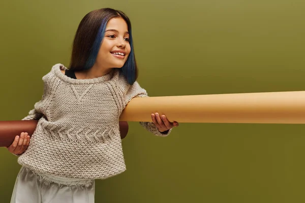 Улыбающаяся и модная девочка-подросток с крашеными волосами в уютном трикотажном свитере, держащая в руках бумагу и отводя взгляд на зеленую, осеннюю моду для детей — стоковое фото