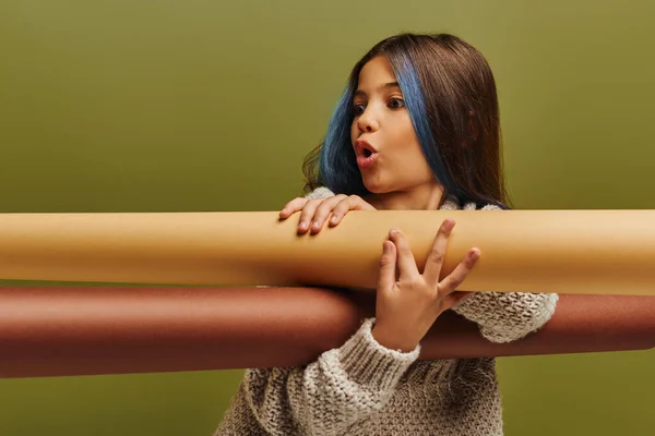 Scioccato preadolescente ragazza con i capelli tinti indossando maglione caldo maglia e tenendo rotoli di carta mentre in piedi e posa isolato su verde, moda autunno per preadolescenti concetto — Foto stock