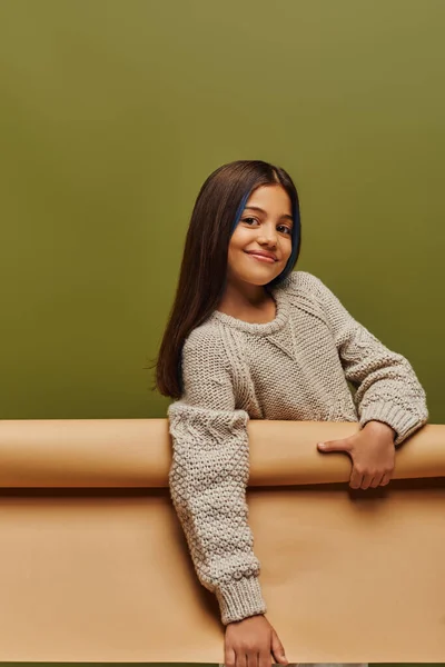Радостная и стильная девочка-подросток с окрашенными волосами в теплый вязаный свитер и смотрящая в камеру, держа в руках бумагу, изолированную на зеленой осенней моде для детей — стоковое фото