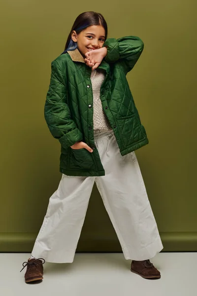 Полная длина позитивной девушки с окрашенными волосами, смотрящей на камеру во время позирования в осеннем наряде и вязаном свитере на зеленом фоне, современная осенняя мода для дошкольников — стоковое фото