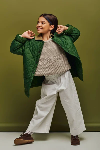Ganzes lächelndes und trendiges Frühchen im Herbst-Outfit und Strickpullover, das wegschaut, während es posiert und auf grünem Hintergrund steht, moderne Kindermode für Frühchen-Konzept — Stockfoto