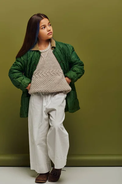 Полная длина модный preadolescent девушка с окрашенными волосами и теплый вязаный свитер, держась за руки на бедрах и позируя на зеленом фоне, современная мода для детей концепции — стоковое фото