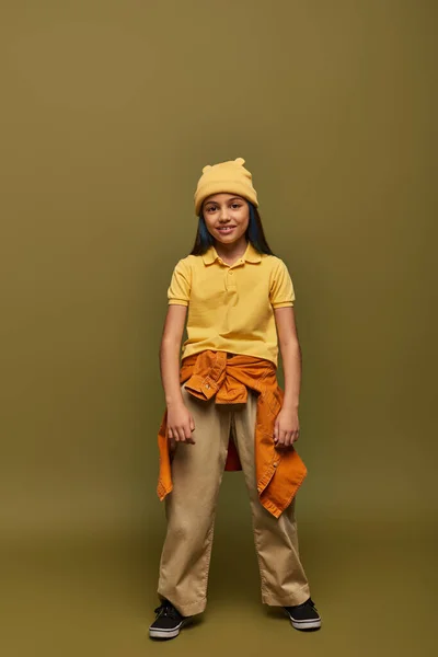 Full length of smiling and stylish preadolescent girl with dyed hair wearing urban outfit and yellow hat while standing on khaki background, stylish girl in modern outfit concept — Stock Photo