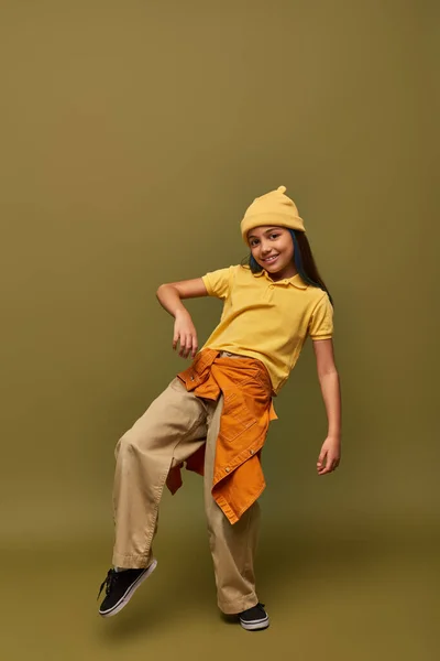 Full length of trendy and cheerful preadolescent girl in urban outfit and yellow hat posing while looking at camera on khaki background, stylish girl in modern outfit concept — Stock Photo