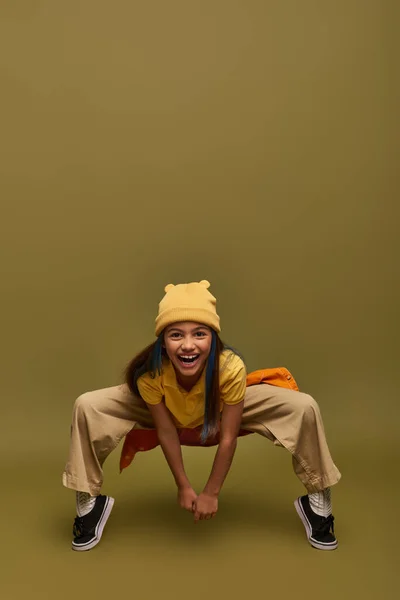 Повна довжина веселої дівчини-підлітка з пофарбованим волоссям в жовтий капелюх і модний одяг позує, дивлячись на камеру на фоні хакі, стильна дівчина в сучасному вбранні концепції — стокове фото