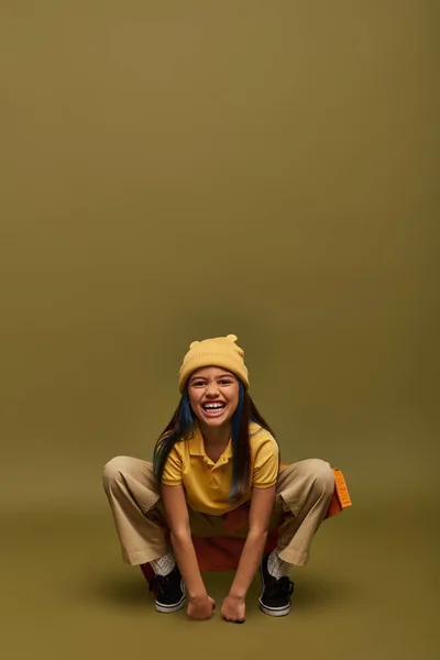 Excited and trendy preteen girl with dyed hair posing in yellow hat and urban outfit while standing and looking at camera on khaki background, stylish girl in modern outfit concept — Stock Photo