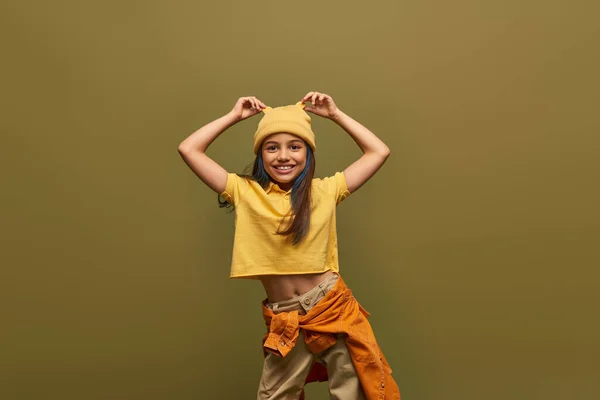 Menina pré-adolescente positivo e elegante com cabelo tingido vestindo roupa urbana e tocando chapéu amarelo enquanto em pé e posando isolado em cáqui, menina elegante no conceito de roupa moderna — Fotografia de Stock