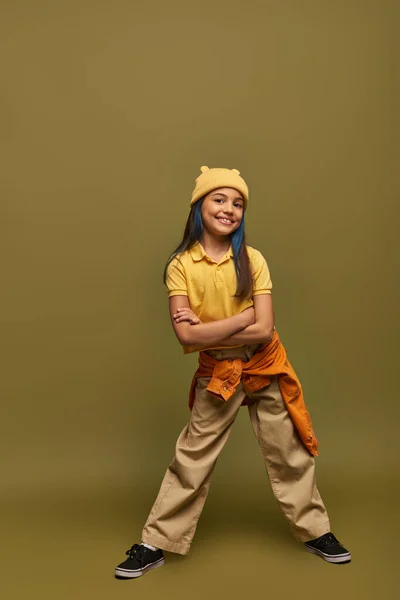 Full length of joyful and stylish preteen girl with dyed hair wearing urban outwear and yellow hat while crossing arms and standing on khaki background, stylish girl in modern outfit concept — Stock Photo