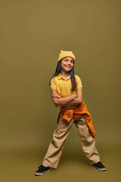 Comprimento total de menina pré-adolescente alegre e elegante com cabelo tingido vestindo roupas urbanas e chapéu amarelo ao cruzar os braços e de pé sobre fundo cáqui, menina elegante no conceito de roupa moderna — Fotografia de Stock