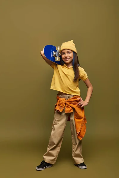 Полная длина веселой и стильной девочки-подростка с окрашенными волосами в городской наряд и желтую шляпу и держа скейтборд на фоне хаки, стильная девушка в современной концепции одежды — стоковое фото