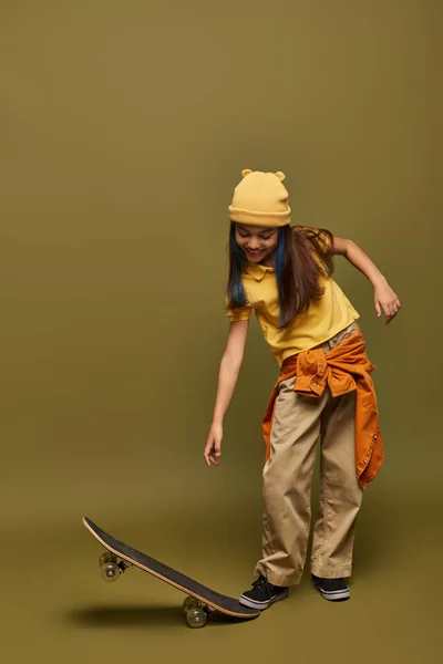 Pleine longueur d'enfant préadolescent joyeux avec des cheveux teints portant chapeau jaune et tenue urbaine tout en regardant skateboard sur fond kaki, fille élégante dans le concept de tenue moderne — Photo de stock