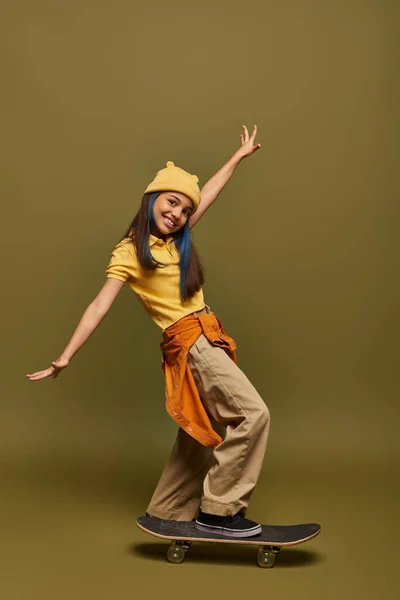 Positive and stylish preadolescent girl with dyed hair wearing yellow hat and urban outfit while looking at camera near skateboard on khaki background, stylish girl in modern outfit concept — Stock Photo