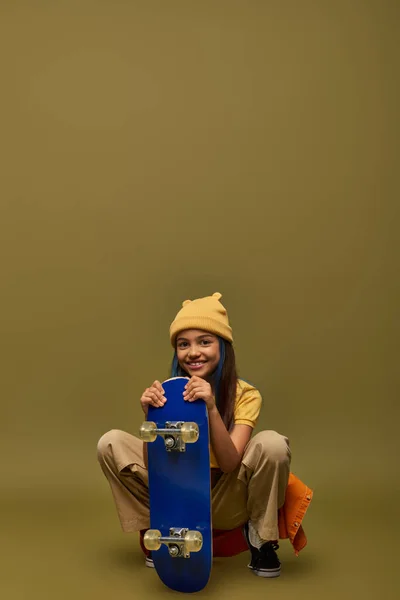 Ragazza alla moda preteen con i capelli tinti indossando cappello giallo e vestito urbano mentre posa con skateboard e seduto su sfondo kaki, ragazza elegante nel concetto di vestito moderno — Foto stock