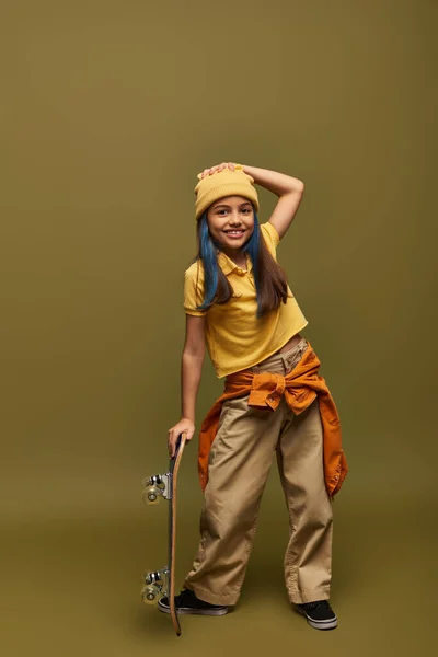 Comprimento total de criança pré-adolescente positiva com cabelo tingido usando chapéu amarelo e roupa urbana e de pé perto de skate no fundo cáqui, menina elegante no conceito de roupa moderna — Fotografia de Stock