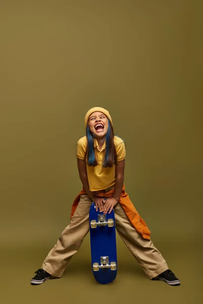 Full length of excited preteen child with colored hair wearing urban outfit and hat while grimacing and holding skateboard on khaki background, stylish girl in modern outfit concept — Stock Photo