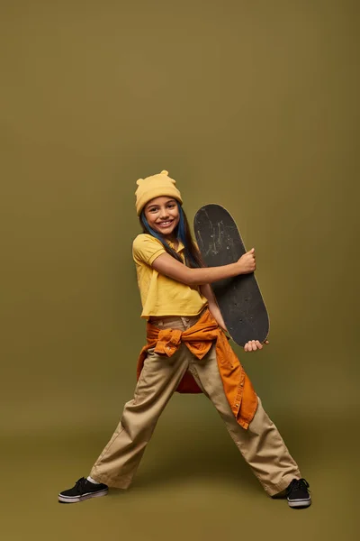 Piena lunghezza di positivo preadolescente ragazza in cappello giallo e vestito urbano in possesso di skateboard e guardando la fotocamera su sfondo kaki, ragazza alla moda nel concetto di abbigliamento moderno — Foto stock