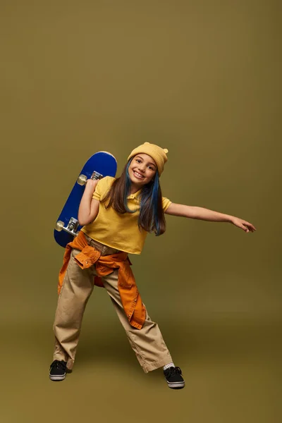 Comprimento total da menina pré-adolescente positiva com cabelo tingido usando chapéu amarelo e roupa urbana enquanto segurando skate e de pé no fundo cáqui, menina elegante no conceito de roupa moderna — Fotografia de Stock