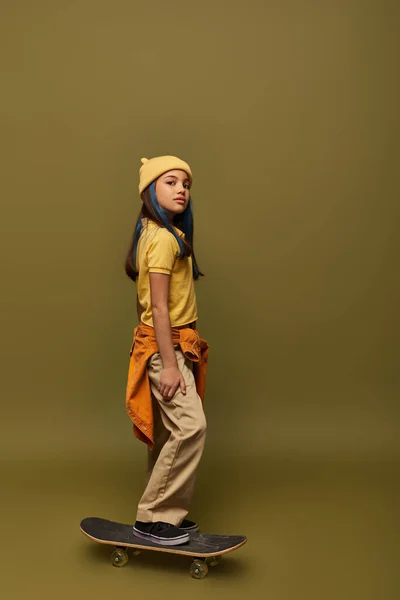 Menina pré-adolescente confiante com cabelo tingido usando chapéu e roupa urbana enquanto olha para a câmera e de pé no skate no fundo cáqui, menina com olhar estilo de rua legal — Fotografia de Stock