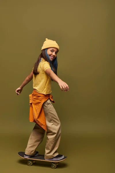 Улыбающаяся и модная девочка-подросток с окрашенными волосами в желтую шляпу и городской наряд, смотрящая в камеру и стоящая на скейтборде на фоне хаки, девушка с прохладным уличным видом — стоковое фото