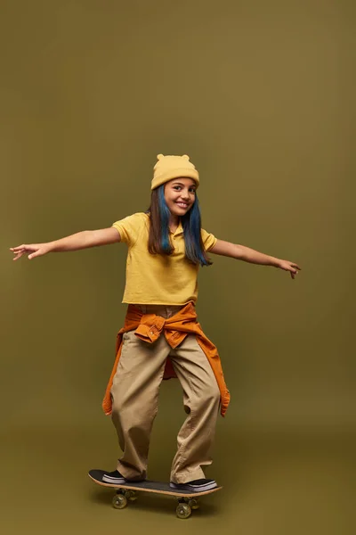 Радостная девочка-подросток с окрашенными волосами в стильный городской наряд и шляпу, стоя на скейтборде и глядя на камеру на фоне хаки, девушка с прохладным уличным видом — стоковое фото