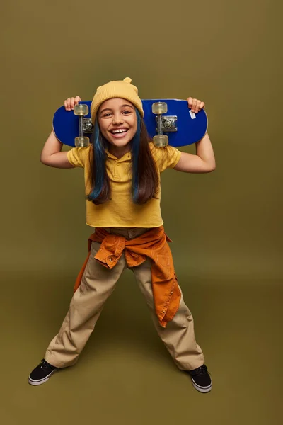 Full length of smiling preteen child with dyed hair wearing yellow hat and urban outfit holding skateboard and looking at camera on khaki background, girl with cool street style look — Stock Photo