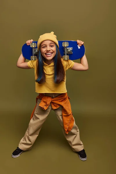 Pleine longueur de préadolescent souriant avec des cheveux teints portant chapeau jaune et tenue urbaine tenant skateboard et regardant la caméra sur fond kaki, fille avec look de style de rue cool — Photo de stock