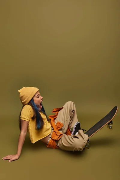 Vista lateral da menina pré-adolescente animado com cabelo colorido vestindo roupa urbana na moda e chapéu enquanto sentado perto de skate no fundo cáqui, menina com olhar estilo de rua legal — Fotografia de Stock