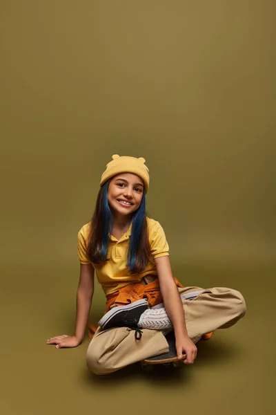 Menina pré-adolescente positiva e elegante com cabelo tingido vestindo roupas urbanas e chapéu amarelo enquanto olha para a câmera e sentado no skate no fundo cáqui, menina com olhar estilo de rua legal — Fotografia de Stock
