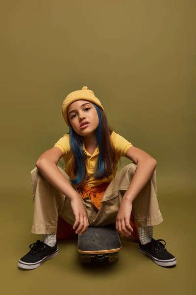 Selbstbewusstes und seriöses Mädchen mit gefärbten Haaren, das urbanes Outfit und Hut trägt, auf Skateboard sitzt und auf khakifarbenem Hintergrund in die Kamera schaut, Mädchen mit coolem Street-Style-Look — Stockfoto