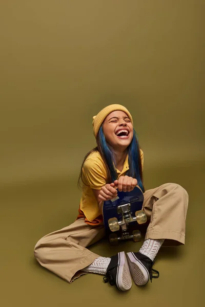 Laughing and stylish preadolescent girl with colored hair wearing urban outfit and yellow hat holding skateboard and sitting on khaki background, girl with cool street style look — Stock Photo
