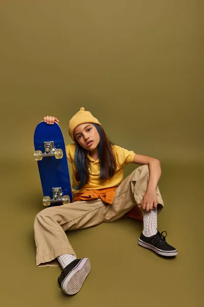 Полная длина уверенного и модного ребенка-подростка с цветными волосами в городской одежде и шляпе, держа скейтборд на фоне хаки, девушка с прохладным уличным видом — стоковое фото