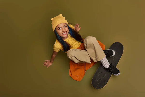 Top view of smiling and stylish preteen girl with colored hair wearing hat and urban outfit while looking at camera and sitting near skateboard on khaki background, girl with cool street style look — Stock Photo