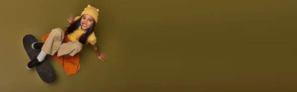 Vista superior da menina pré-adolescente na moda em chapéu e roupas urbanas elegantes sorrindo para a câmera enquanto sentado perto de skate no fundo cáqui, menina com olhar estilo de rua legal, banner — Fotografia de Stock