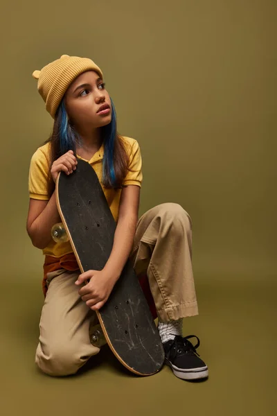 Portrait of trendy preadolescent girl with colored hair wearing yellow hat and urban outfit while holding skateboard and looking away on khaki background, girl with cool street style look — Stock Photo